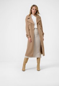 Tan double-breasted coat with wide collar, beige textured skirt, and light beige knee-high boots in a minimalistic setting.