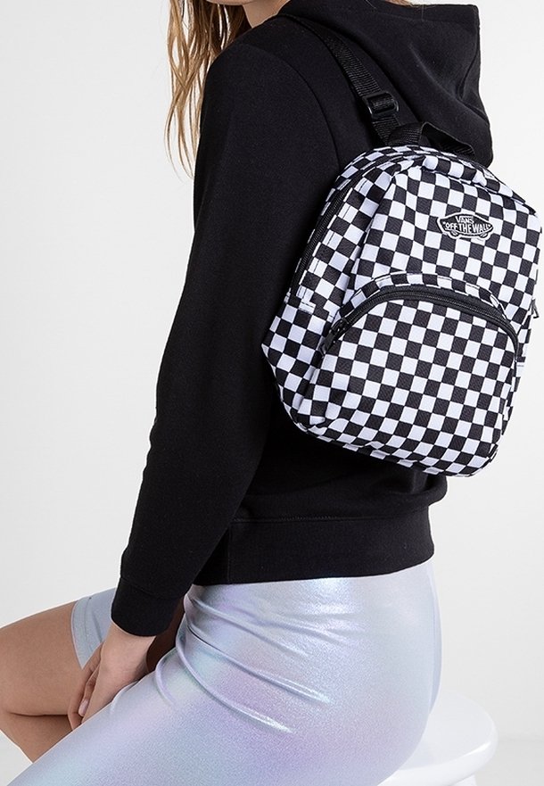 Personne portant un sweat à capuche noir et une jupe argentée brillante assise avec un sac à dos Vans à carreaux noir et blanc sur l’épaule.
