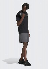 Svart kortermet treningstrøye med logo, kombinert med grå shorts. Modellen har på seg svarte treningssko og viser en sporty holdning.