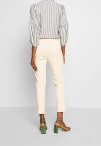 Kvinna som bär krämfärgade cropped byxor, en grå och vit randig blus med ballongsärmar, samt gröna högklackade sandaletter med remmar, stående.