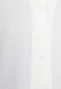 Camisa branca com colarinho, feita de um tecido suave e leve. Apresenta botões brancos simples e uma textura discreta, exibindo um design clássico.
