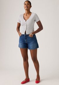 Γυναίκα που στέκεται με τα χέρια στις τσέπες, φορώντας ριγέ κοντομάνικη μπλούζα, τζιν σορτς και κόκκινα επίπεδα παπούτσια μπροστά σε ουδέτερο φόντο.