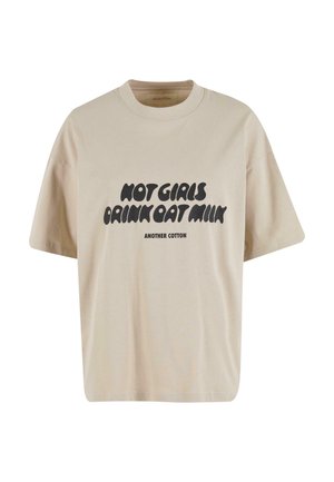 Camiseta de algodón beige con un corte holgado, escote redondo y mangas cortas. Presenta un texto gráfico en negro: "LAS CHICAS CALIENTES BEBEN LECHE DE AVENA."