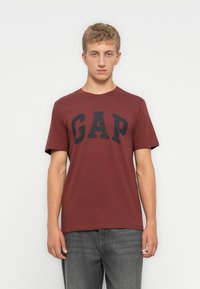 Ung mand iført en kastanjefarvet T-shirt med korte ærmer og sort "GAP" logo samt grå jeans, stående mod en ensfarvet hvid baggrund.