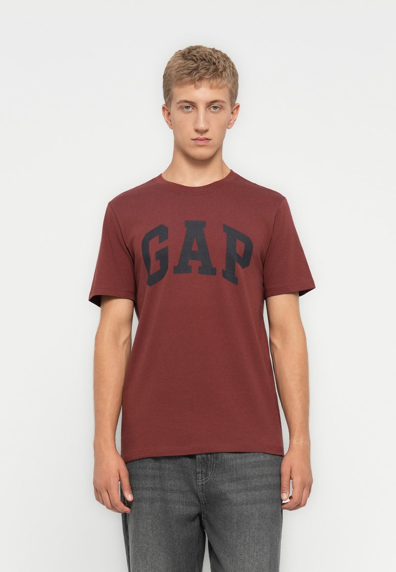Ung mand iført en kastanjefarvet T-shirt med korte ærmer og sort "GAP" logo samt grå jeans, stående mod en ensfarvet hvid baggrund.