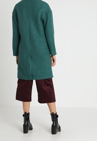 Cappotto di lana teal con taglio dritto e spacco sul retro, abbinato a culotte in velluto a costine bordeaux e stivali neri alla caviglia con tacco grosso.