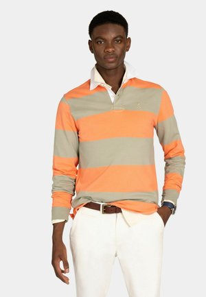 Joven con una camiseta de rugby de manga larga con anchas franjas horizontales naranjas y beige, pantalones blancos, cinturón marrón y reloj de pulsera.