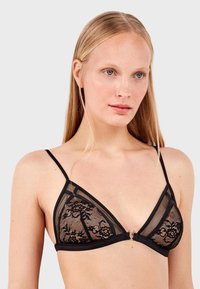 Bralette triangular de encaje negro con paneles de malla transparente, patrones florales, tirantes finos y un pequeño conector de metal dorado en la parte delantera.