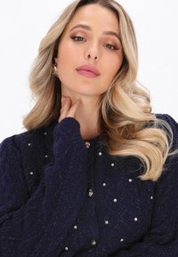 Tummansininen neulecardigan, jossa on kabelikuvio, metallinapit ja etuosassa kimalteleva strassikoristus. Pitkät, teksturoidut hihat.