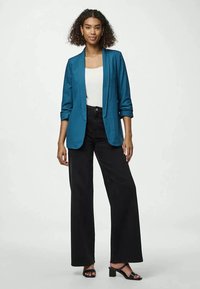 Blazer teal com um design sob medida, combinado com uma blusa branca e calças largas pretas de cintura alta. Sandálias de salto bloco pretas.