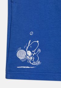 A kék textilen egy fehér illusztráció található, amelyen egy mesefigura kutya teniszmezőn játszik, ütőt tart a mancsában, és egy labda lebeg a feje fölött. Sima felület, szoros kötött minta.