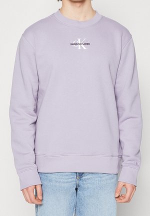 Hombre con sudadera color púrpura claro de Calvin Klein Jeans con el logo en el pecho y la manga, combinado con jeans azul claro, posando contra un fondo blanco.