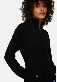 Sweat à fermeture éclair noir avec un col côtelé et des manches longues, présenté dans un tissu tricoté texturé et un design ajusté.