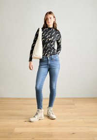 Haut noir à manches longues avec un motif abstrait beige, jeans skinny bleus et bottines beige clair à la cheville. La personne tient un sac à bandoulière beige.