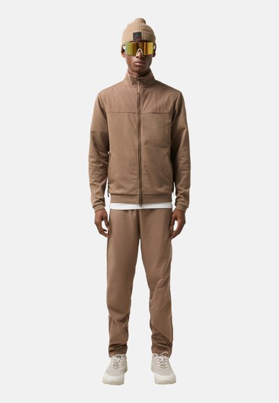 Brown athletic jacket with high collar, matching pants, and a beige knit beanie. Reflective sunglasses with yellow lenses complete the look.