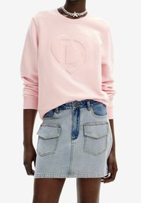 Felpa rosa con cuori in rilievo e logo 'D', abbinata a una gonna in denim chiaro con due tasche frontali e una toppa blu.