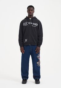 Felpa nera con zip e logo grigio Ed Hardy, abbinata a jeans in denim blu con ricamo colorato di drago sulla gamba sinistra. Sneakers nere.