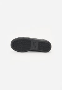 Fekete sneaker talp, textúrált cikk-cakk mintával, gumiból készült, és egy téglalap alakú márkacímke látható a boltív területén.