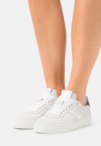 Białe zamszowe sneakersy o fakturowanym wykończeniu, z czarnym skórzanym akcentem na pięcie, okrągłym noskiem oraz białymi sznurowadłami na podwyższonej podeszwie.