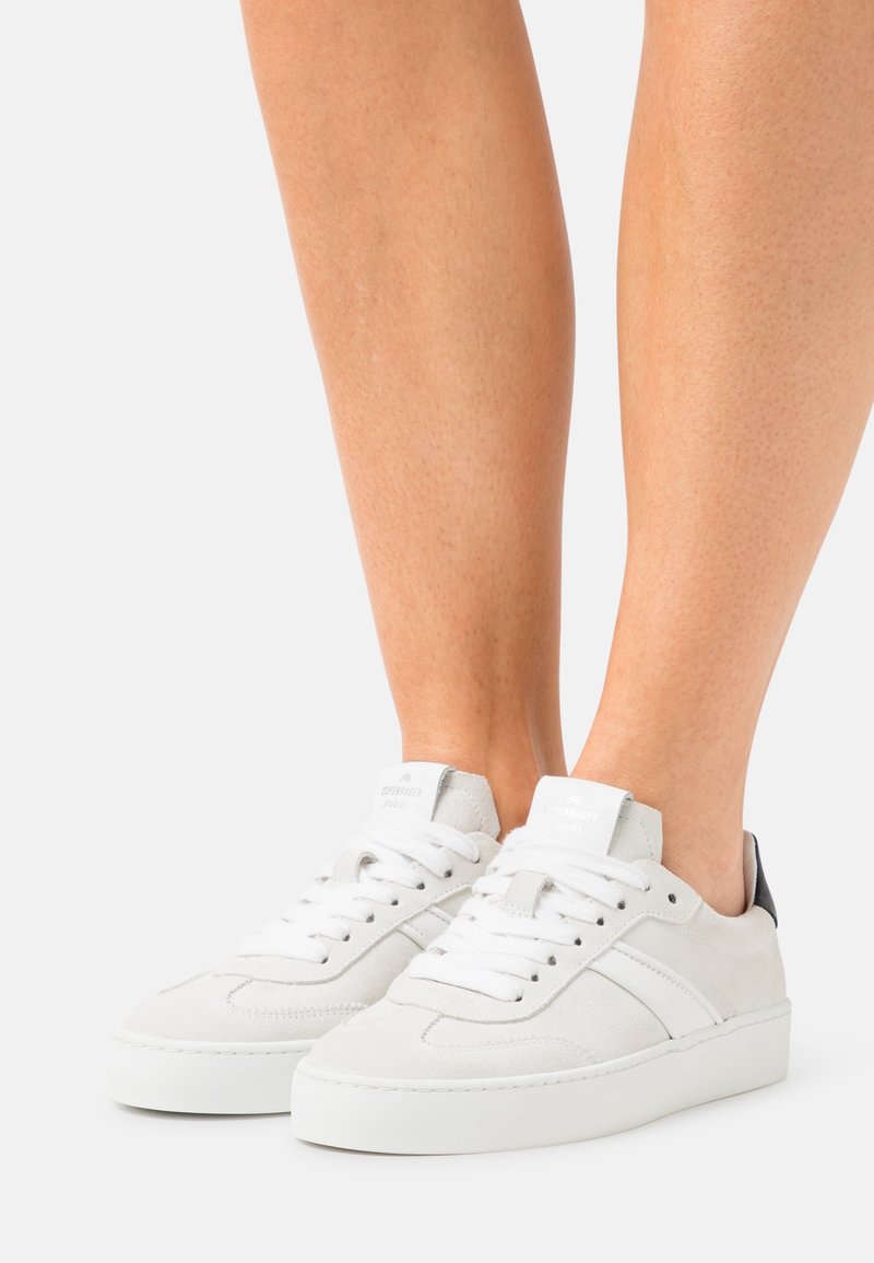 Białe zamszowe sneakersy o fakturowanym wykończeniu, z czarnym skórzanym akcentem na pięcie, okrągłym noskiem oraz białymi sznurowadłami na podwyższonej podeszwie.