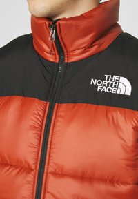 Rote und schwarze Daunenjacke mit hohem Kragen, ausgestattet mit einem Reißverschluss vorne und einem Logo auf der oberen linken Seite. Strukturierter, gesteppten Design.