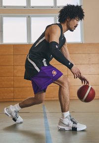 Moški košarkar dribla rdečo in belo žogo, oblečen v črno majico, vijolične kratke hlače in bele superge Nike z sivo barvo.