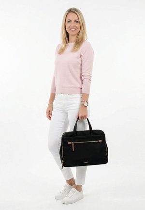 Femme portant un pull rose et un pantalon blanc tenant un sac à main noir avec une poche zippée à l'avant, debout sur un fond blanc.