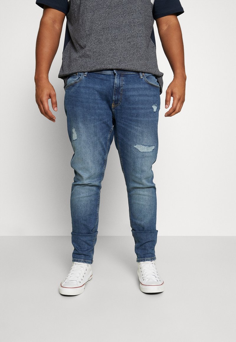 Jeans en denim bleu délavé à la coupe slim, avec des zones usées et un ourlet retroussé. Associés à des baskets blanches.