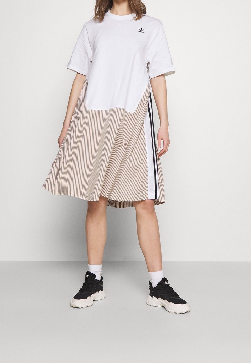 Valkoinen T-paitamekko, jossa on vaakasuuntaiset beigealareunat, mustat sivuviivat ja logo vasemmalla ylhäällä. Yhdistetty mustiin lenkkareihin.