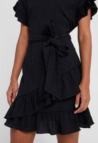 Schwarzes Kleid aus strukturiertem Stoff mit Rüschensäumen, gebundenem Taillengürtel und mehrschichtigen Rüschendetails am Saum.