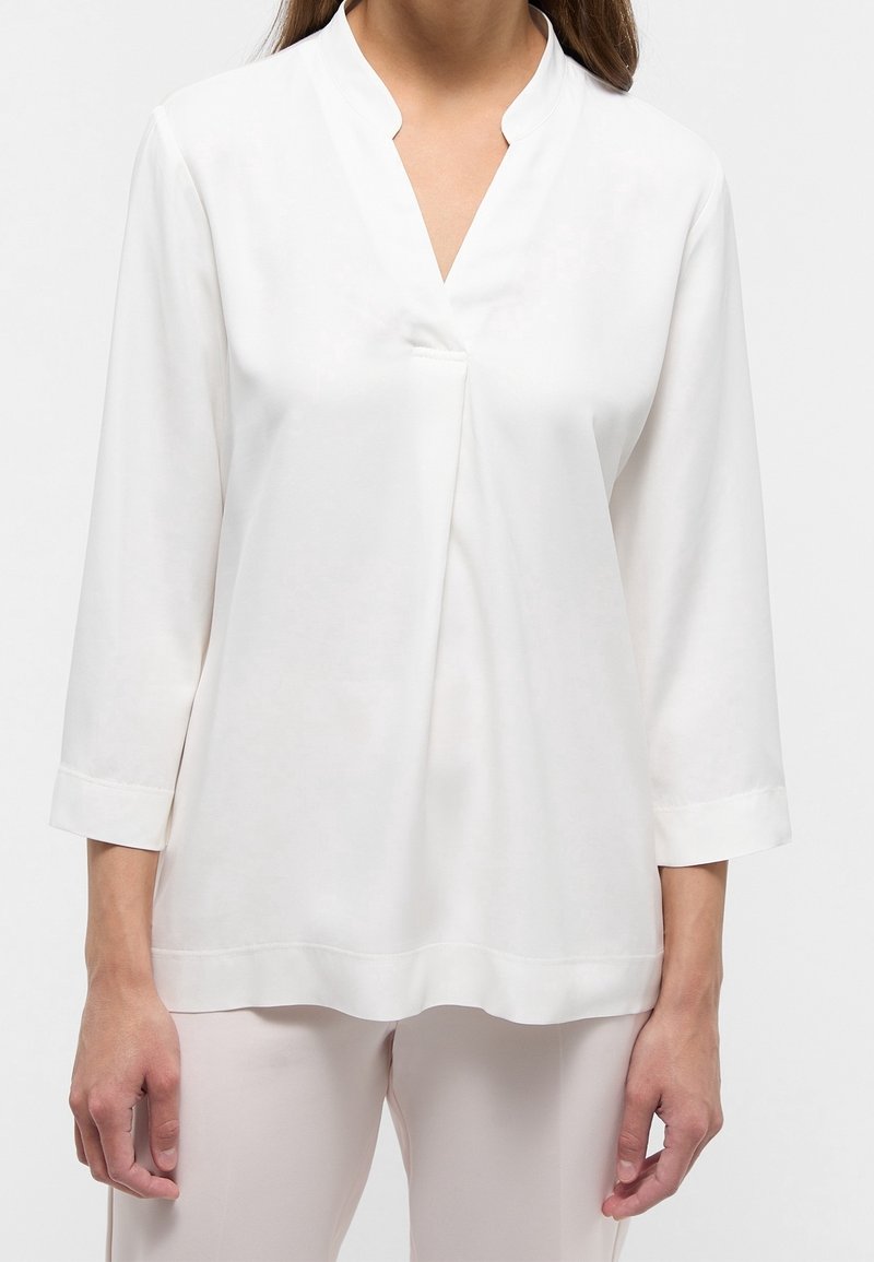 Blusa bianca con maniche lunghe e scollo a V, caratterizzata da un sottile dettaglio plissettato sul davanti e un orlo dritto con accenti cuciti.