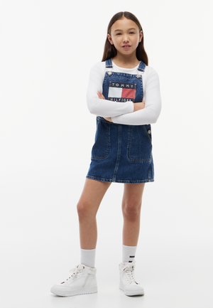 Chica de pie con confianza, con los brazos cruzados, vistiendo una camisa blanca de manga larga bajo un vestido peto de mezclilla de Tommy Hilfiger y zapatillas blancas.