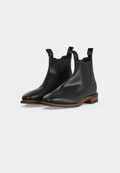 Paire de bottines Chelsea en cuir noir avec des panneaux latéraux élastiques et des semelles en bois marron sur fond clair.