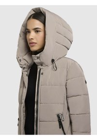 Giacca puffer beige chiaro con un grande cappuccio foderato, texture trapuntata, zip frontale e tasche laterali, con dettagli in metallo nero.