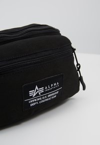 Svart stoffpose med minst to glidelåslommer, som har et merke med "Alpha Industries" og teksten "Official U.S. Defense".