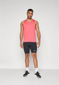 Nike sleeveless pink athletic shirt with logo, paired with black shorts, and black running shoes. Model has short hair and stands upright.