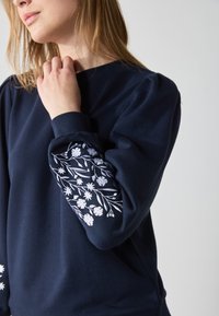 Námornícky modrý sweatshirt s dlhými rukávmi, ktorý obsahuje biele kvetinové vyšívanie na ľavom rukáve a jemnú, štruktúrovanú tkaninu.