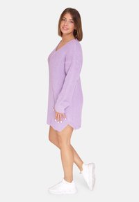 Lavendelfarbenes, oversized gestricktes Kleid mit V-Ausschnitt, langen Ärmeln und einem Saum mit seitlichen Schlitzen, kombiniert mit weißen Sportschuhen.