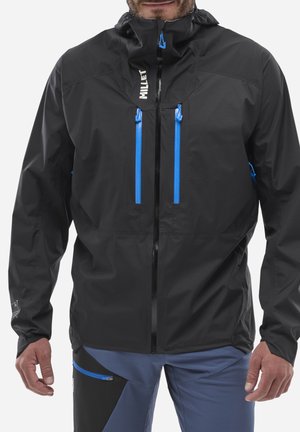 Homme portant une veste imperméable noire avec des fermetures éclair bleues et un pantalon de plein air bleu et noir, debout les bras détendus.