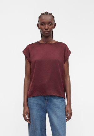 Femme aux cheveux tressés portant un haut bordeaux sans manches et un jean bleu, debout devant un fond blanc uni.