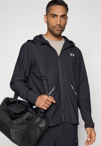 Musta vetoketjullinen takki hupulla, jossa valkoiset yksityiskohdat ja joustavaa materiaalia. Malli pitää mustaa treenikassia, jossa on hihna.