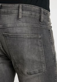 Jeans de mezclilla gris con un clásico diseño de cinco bolsillos y una textura sutil, con un bolsillo trasero visible y detalles en la cinturilla.
