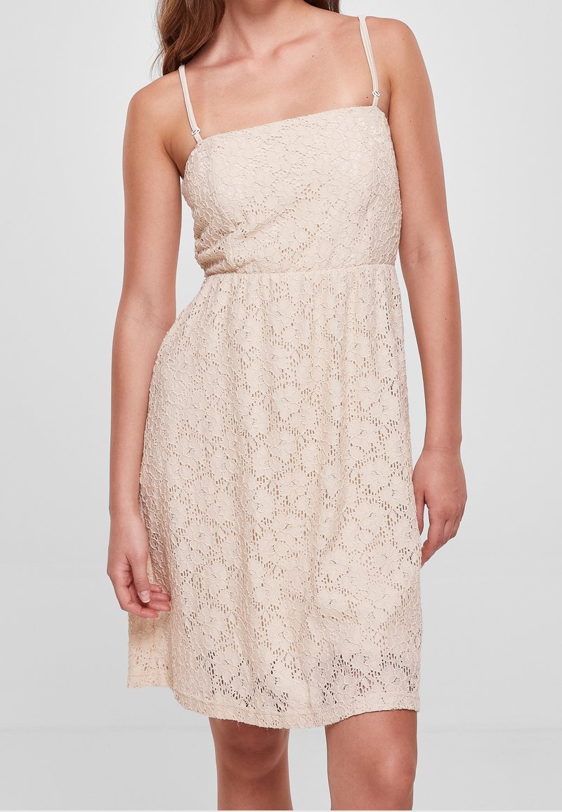 Femme portant une robe en dentelle beige mi-longue avec de fines bretelles spaghetti réglables et un corsage cintré.