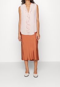 Blouse sans manches rose clair avec un devant à volants, associée à une jupe en satin rouille arrivant au genou et des chaussures blanches à talons blocs.