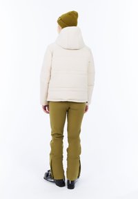 Persoon in een crèmekleurige pufferjack, olijfgroene broek, olijfgroene gebreide muts en zwarte skibotten, staande tegen een witte achtergrond.