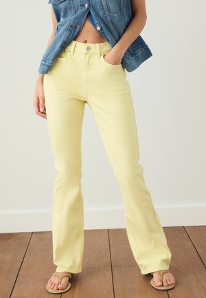 Femme portant un jean taille haute jaune clair, une veste en jean bleue et des tongs beiges, debout sur un sol en bois contre un mur blanc.