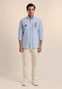 Camisa de botones azul claro con dos insignias y mangas largas, combinada con pantalones cargo crema y zapatillas blancas. Textura de tela suave.