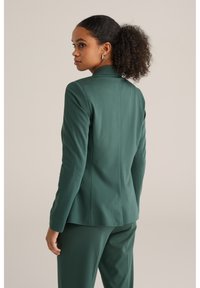 Zelený oblekový blejzer so slim fit strihom, jedným gombíkom a hladkou textúrou, skombinovaný so zodpovedajúcimi nohavicami. Zadný pohľad ukazuje detaily švu.