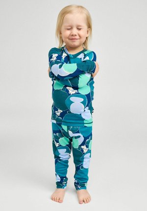 Bambino piccolo con gli occhi chiusi, che si abbraccia, indossa una maglietta e pantaloni con fantasia blu-verde, in piedi a piedi nudi su sfondo chiaro.