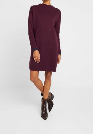 Femme portant une robe prune à manches longues, longueur genou, avec des poignets noirs et des bottines à motif peau de serpent à bout pointu.